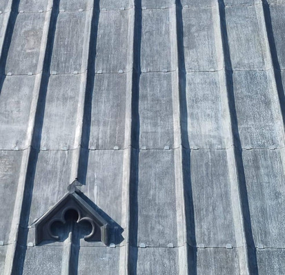 Ein graues Metalldach mit vertikalen Nähten und einem kleinen, dekorativen dreieckigen Fenster unten links. - © Groupe Balas