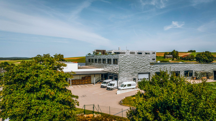 Modernes Industriegebäude mit strukturierter Fassade, umgeben von Bäumen und geparkten weißen Lieferwagen unter blauem Himmel. - © Bild: Kraiss