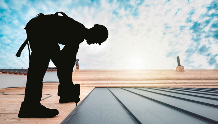 Silhouette eines Arbeiters, der ein Metalldach auf einem Gebäude unter einem hellen, teilweise bewölkten Himmel installiert. - © Bild: Sabrewolf - stock.adobe.com
