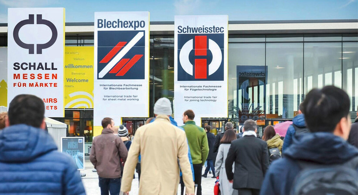 Menschen gehen auf den Eingang eines Kongresszentrums zu, über dem große Messebanner hängen. - © Bild: P. E. Schall GmbH & Co. KG